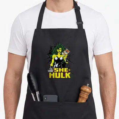 Comic savage apron - She-Hulk holding comic book with stacked text and cracked black background — shown on a black cotton canvas apron against a white background