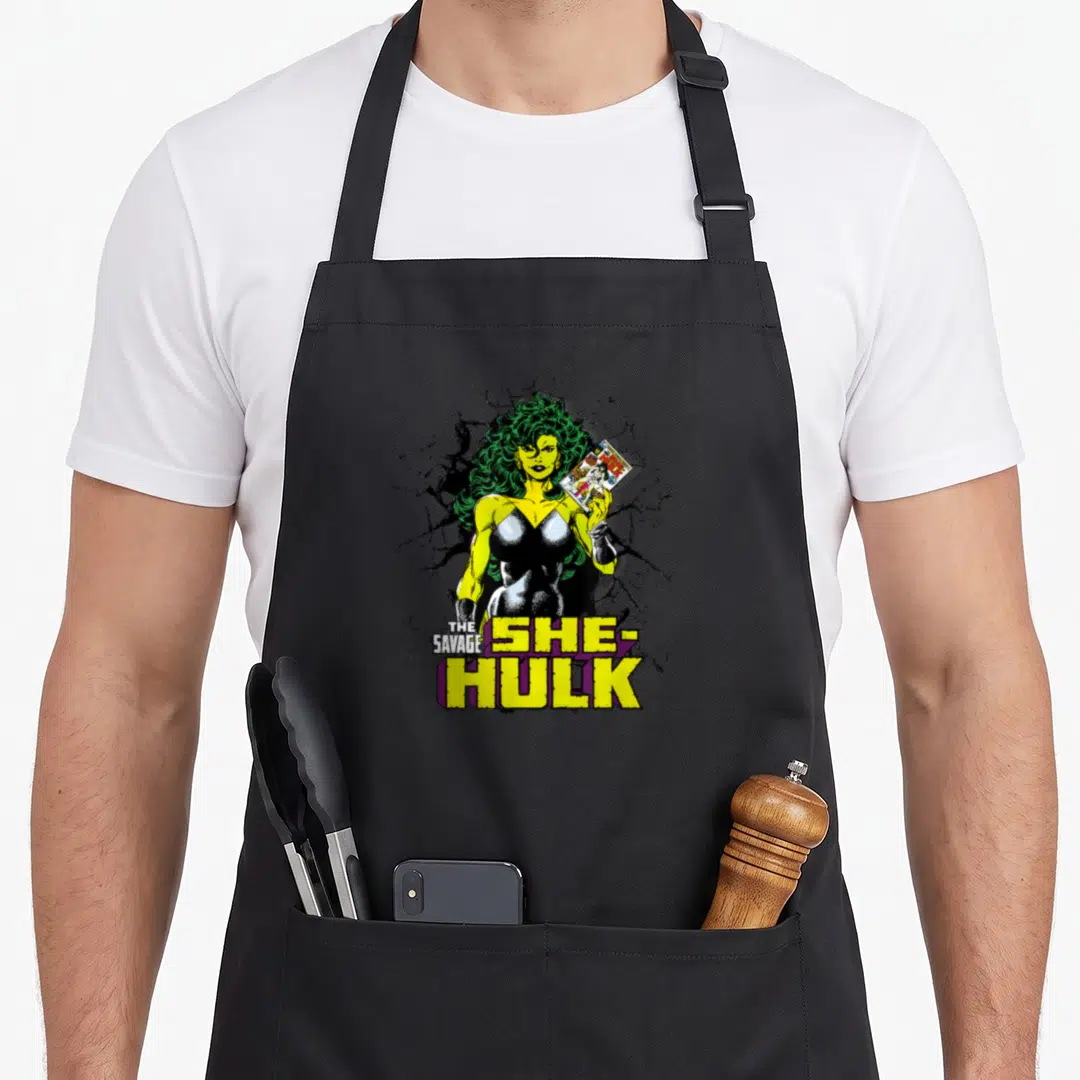 Comic savage apron - She-Hulk holding comic book with stacked text and cracked black background — shown on a black cotton canvas apron against a white background