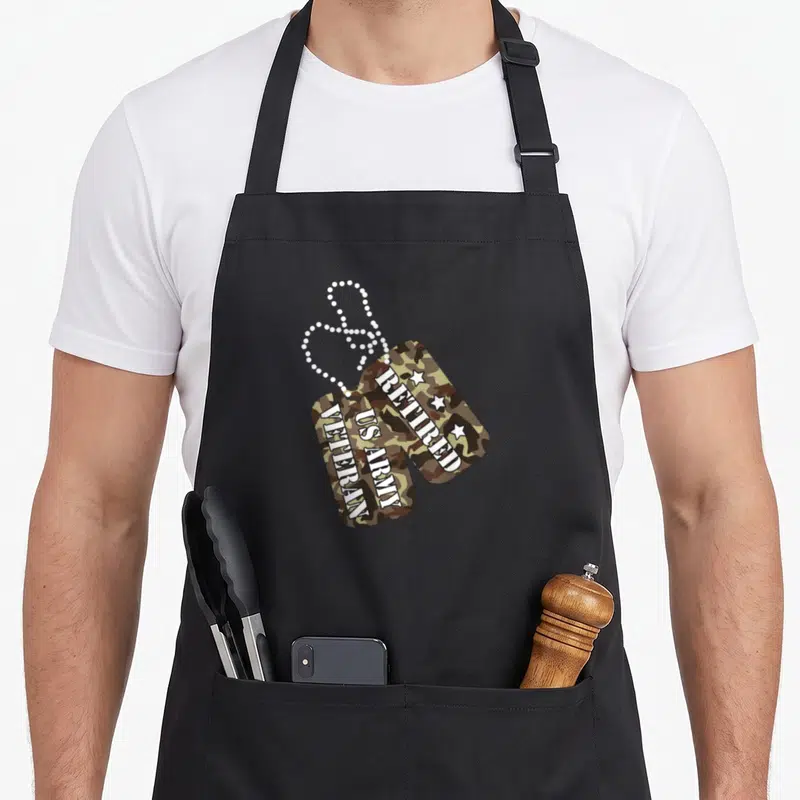 Retired dog apron - Two camo dog tags with vertical RETIRED US ARMY VETERAN text and white stars on a chain — shown on a black cotton canvas apron against a white background