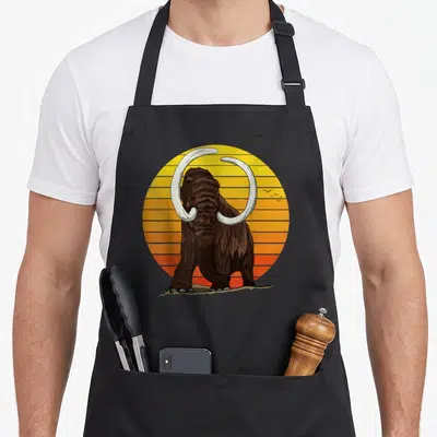 Woolly mammoth apron - Brown mammoth with curved tusks in front of striped yellow orange sun with three birds and grass — shown on a black cotton canvas apron against a white background