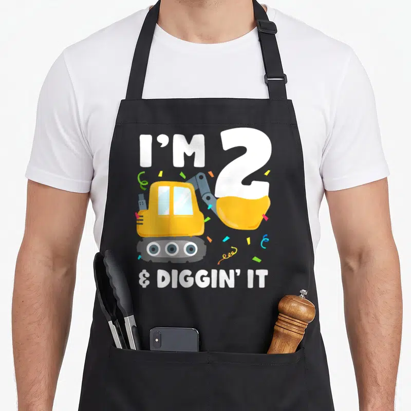 Construction 2nd birthday truck apron - Yellow excavator with I'M 2 & DIGGIN IT text and scattered confetti in cartoon — shown on a black cotton canvas apron against a white background