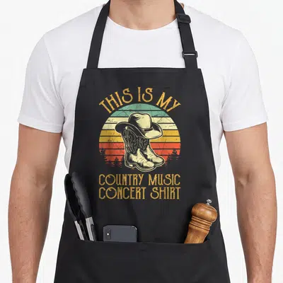 Music country kitchen apron - Retro cowboy boots and hat over striped sun with text THIS IS MY COUNTRY MUSIC CONCERT — shown on a black cotton canvas apron against a white background
