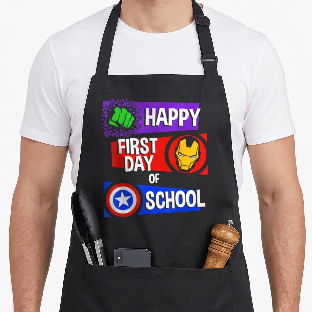 Funny school apron - HAPPY FIRST DAY OF SCHOOL text with fist, helmet, and shield on purple red blue banners — shown on a black cotton canvas apron against a white background