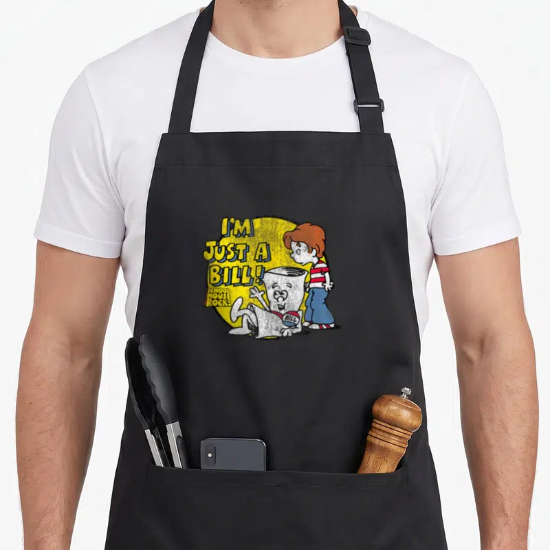 Rock show apron - I'm Just a Bill text in yellow bubble with cartoon boy and paper character — shown on a black cotton canvas apron against a white background
