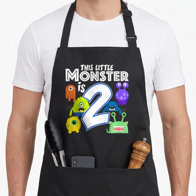 Monster 2nd apron - Five colorful cartoon monsters surrounding a large number 2 with stacked white text — shown on a black cotton canvas apron against a white background