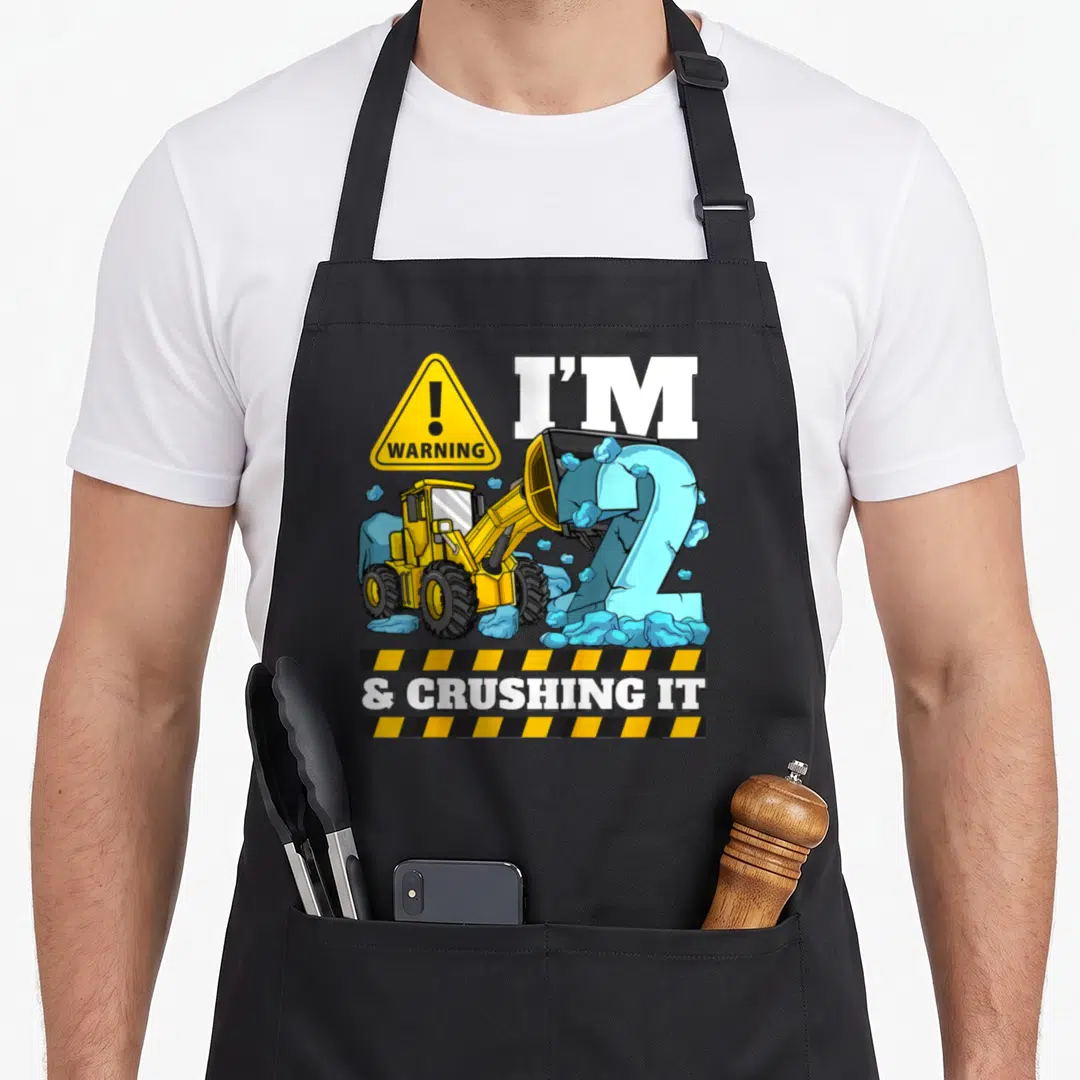 Birthday bulldozer truck apron - Yellow front loader crushing blue number 2 with warning sign and hazard stripe text — shown on a black cotton canvas apron against a white background