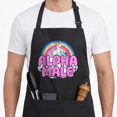 Ironic alpha apron - Cartoon unicorn with pink mane under rainbow and bold pink ALPHA MALE text — shown on a black cotton canvas apron against a white background
