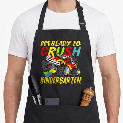 Monster school truck apron - I'M READY TO CRUSH KINDERGARTEN text with cartoon monster truck and colorful splatter — shown on a black cotton canvas apron against a white background