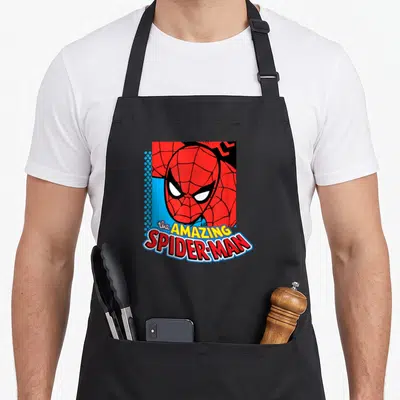 Amazing essential apron - Cartoon Spider-Man mask in a square frame with stacked red and yellow logo text below — shown on a black cotton canvas apron against a white background