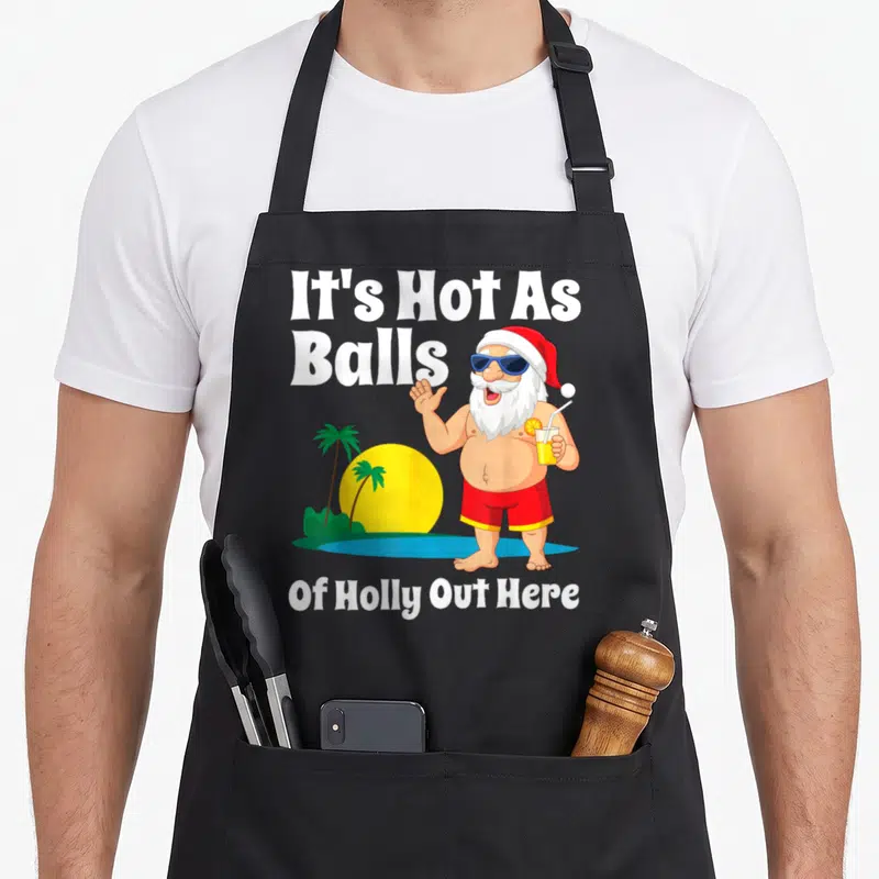 Hot christmas santa apron - Cartoon Santa with sunglasses holding a drink next to palm trees and text It's Hot As Balls — shown on a black cotton canvas apron against a white background