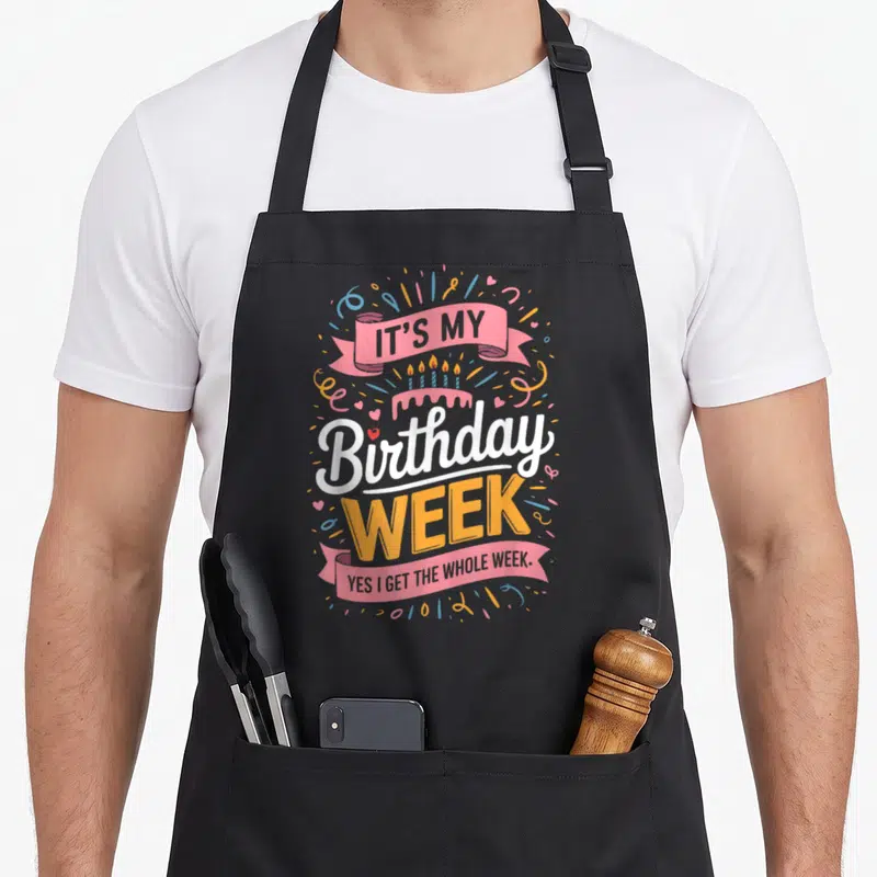 Cool week birthday apron - Cartoon cake with pink ribbon and text IT'S MY Birthday WEEK YES I GET THE WHOLE WEEK — shown on a black cotton canvas apron against a white background