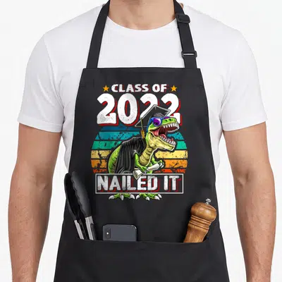 Gown dinosaur apron - T-Rex in cap gown sunglasses holding diploma with Class of 2022 Nailed It text — shown on a black cotton canvas apron against a white background
