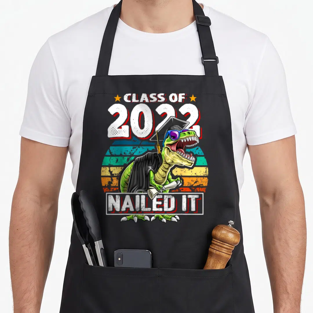 Gown dinosaur apron - T-Rex in cap gown sunglasses holding diploma with Class of 2022 Nailed It text — shown on a black cotton canvas apron against a white background