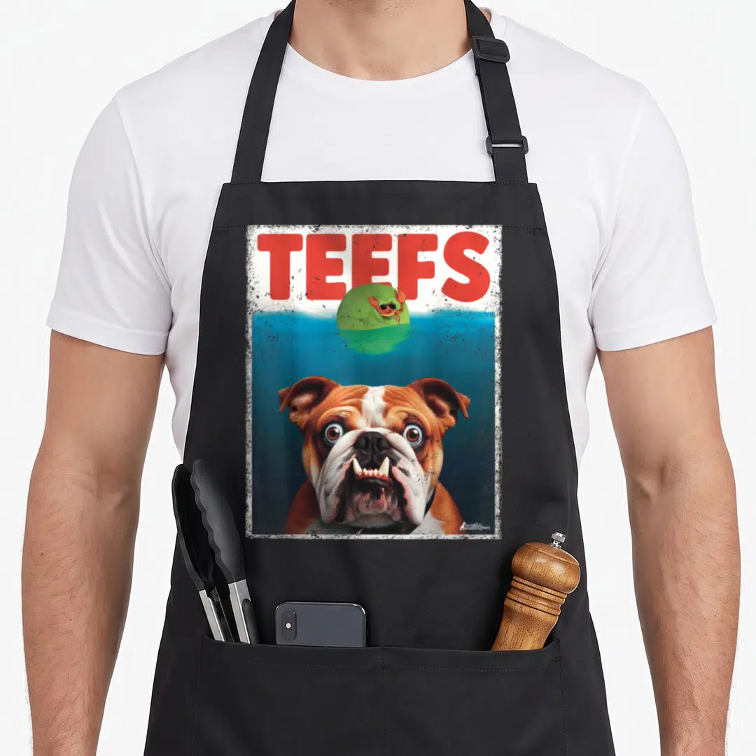 Teef english aprons - Cartoon bulldog head in blue water below red TEEFS text and green island with crab — shown on a black cotton canvas apron against a white background
