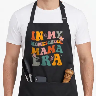 Homeschool teacher apron - Retro IN MY HOMESCHOOL MAMA ERA text with butterflies flowers and sparkles in orange pink — shown on a black cotton canvas apron against a white background