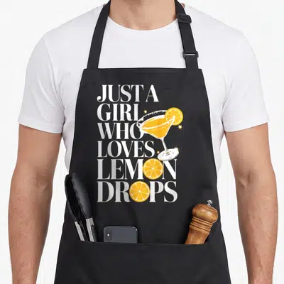 Cocktail for love and lemons apron - Stacked white text reads Just A Girl Who Loves Lemon Drops beside a yellow martini — shown on a black cotton canvas apron against a white background