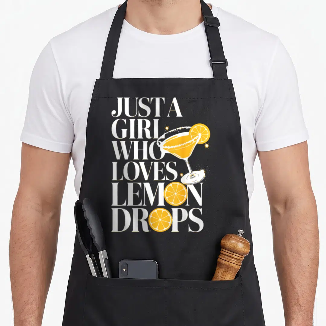 Cocktail for love and lemons apron - Stacked white text reads Just A Girl Who Loves Lemon Drops beside a yellow martini — shown on a black cotton canvas apron against a white background