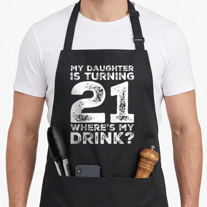 21st mom daughter apron - White distressed text reading MY DAUGHTER IS TURNING 21 WHERE'S MY DRINK — shown on a black cotton canvas apron against a white background