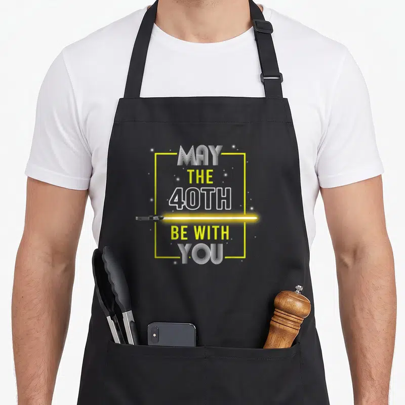 Fortieth 40th birthday apron - Yellow lightsaber separates stacked white text reading May The 40th Be With You inside — shown on a black cotton canvas apron against a white background