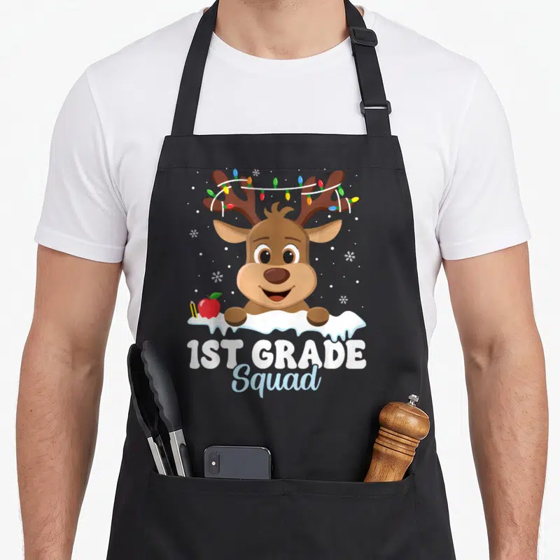 1st christmas aprons - Cartoon reindeer with antler lights, apple, pencil, snowbank, and 1ST GRADE Squad text — shown on a black cotton canvas apron against a white background