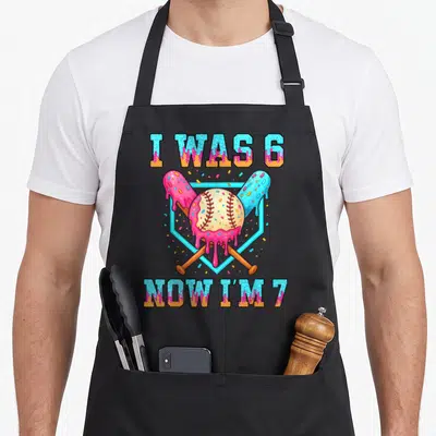 7th ice cream apron - I WAS 6 NOW I'M 7 text over pink icing baseball and crossed bats in blue hexagon — shown on a black cotton canvas apron against a white background