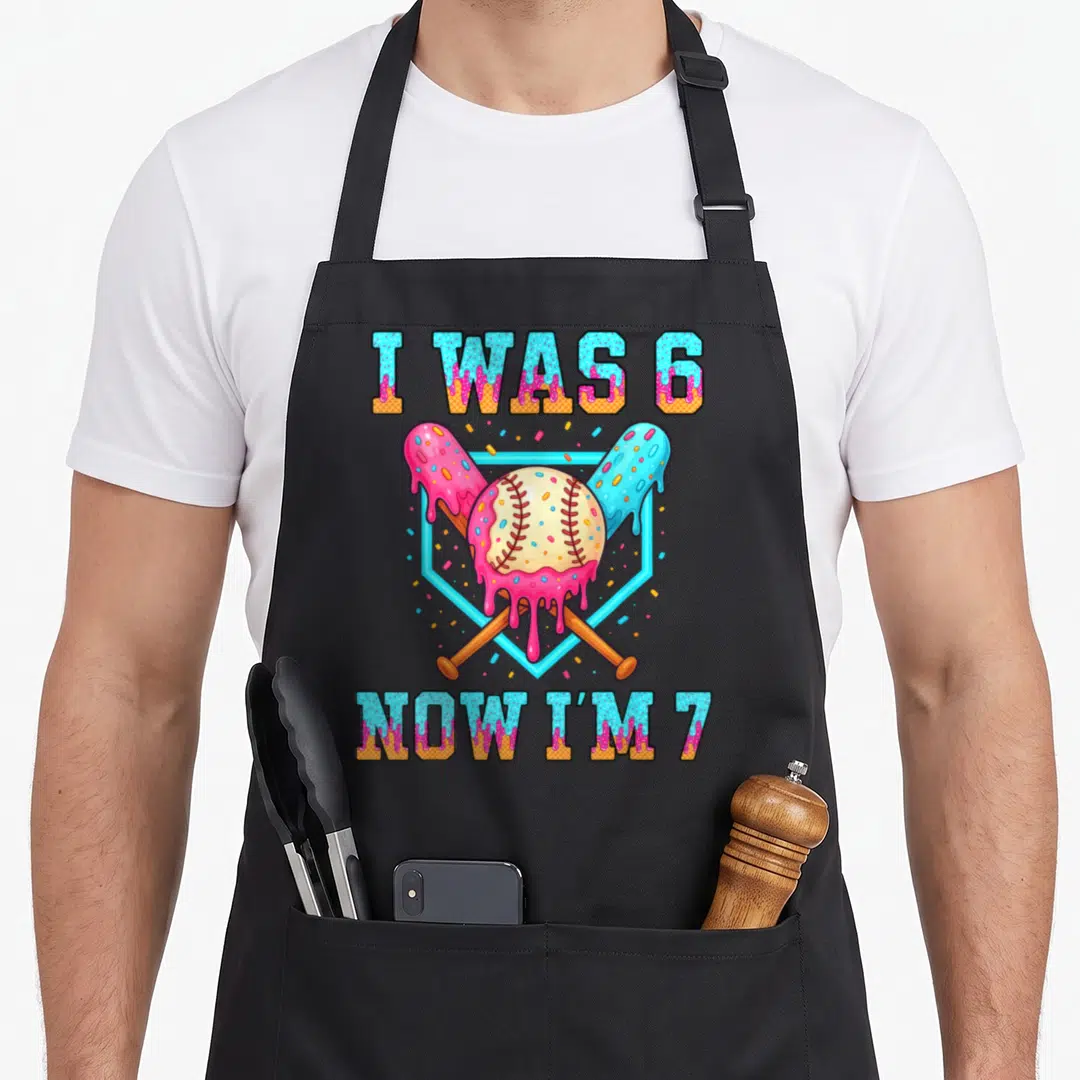 7th ice cream apron - I WAS 6 NOW I'M 7 text over pink icing baseball and crossed bats in blue hexagon — shown on a black cotton canvas apron against a white background