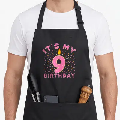 9th donut apron - Pink doughnut letters IT'S MY BIRTHDAY surrounding a number 9 candle with sprinkles — shown on a black cotton canvas apron against a white background