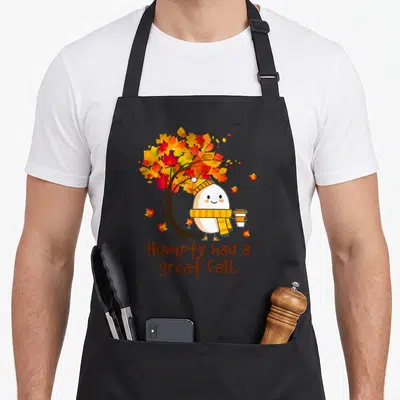 Vintage humpty cute fall aprons - Cartoon egg character with scarf under orange and yellow tree with brown text — shown on a black cotton canvas apron against a white background
