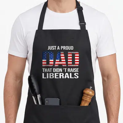 Proud novelty apron - Distressed text Just a Proud Dad That Didn't Raise Liberals with flag pattern fill in DAD — shown on a black cotton canvas apron against a white background