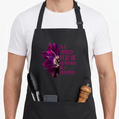 Anemone grandma's aprons - Purple anemone flower next to stacked pink text reading IN A WORLD FULL OF GRANDMAS BE — shown on a black cotton canvas apron against a white background