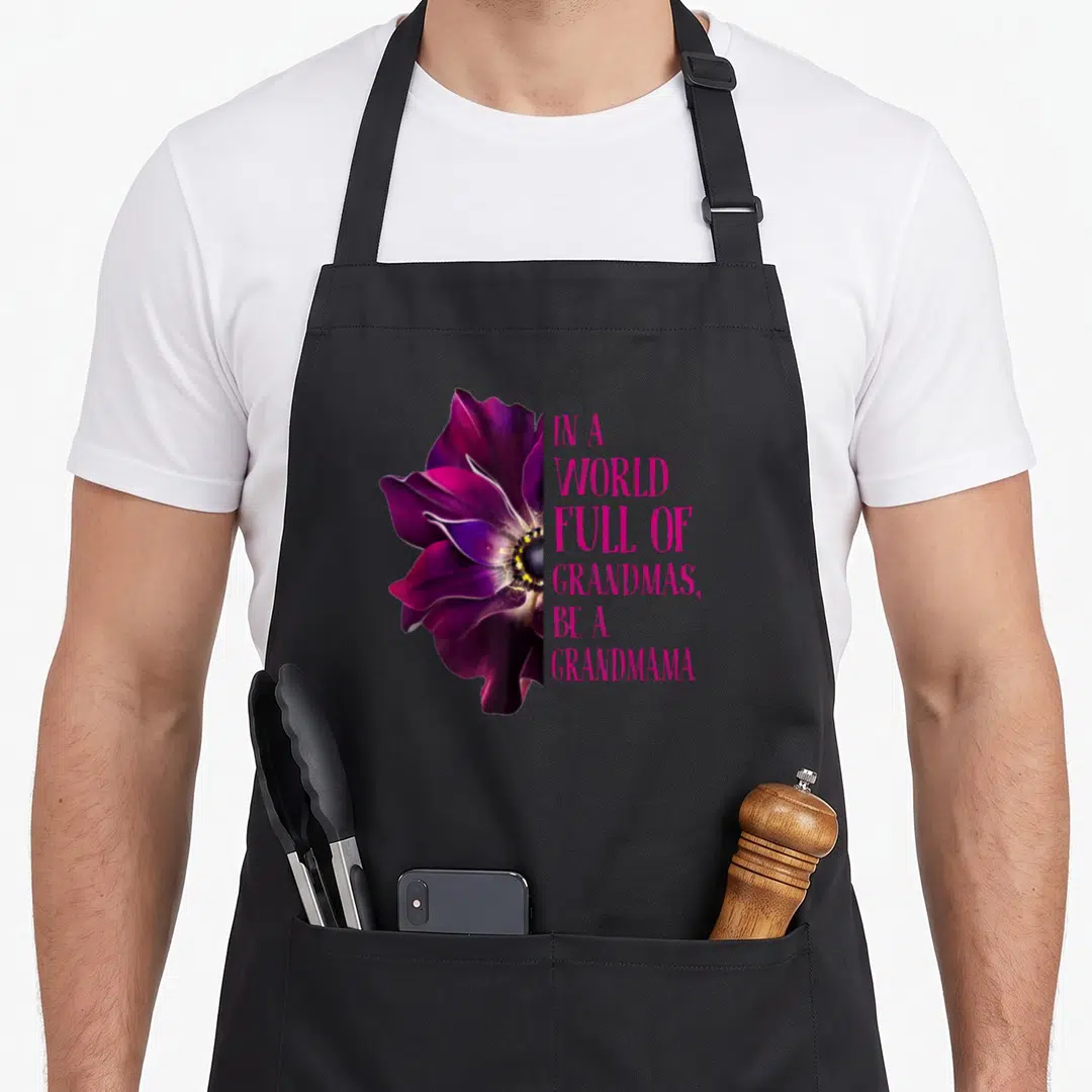 Anemone grandma's aprons - Purple anemone flower next to stacked pink text reading IN A WORLD FULL OF GRANDMAS BE — shown on a black cotton canvas apron against a white background