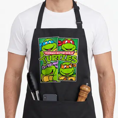 Character teenage cooking aprons - Four smiling turtles in blue red purple orange masks with TEENAGE MUTANT NINJA — shown on a black cotton canvas apron against a white background