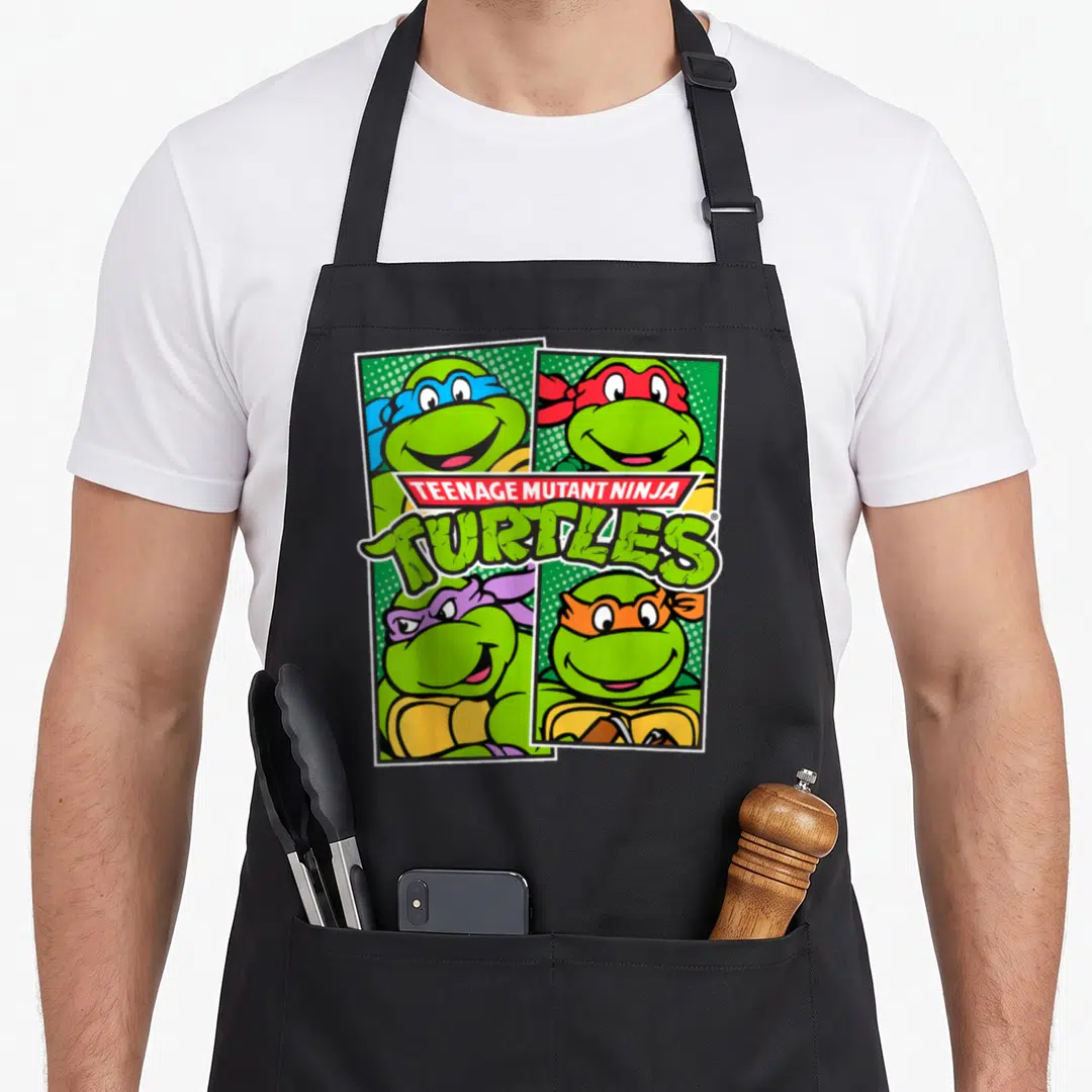 Character teenage cooking aprons - Four smiling turtles in blue red purple orange masks with TEENAGE MUTANT NINJA — shown on a black cotton canvas apron against a white background