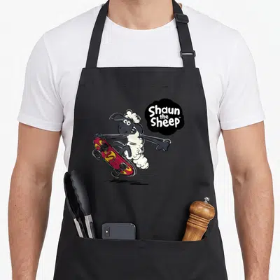 Skateboarding sheep apron - Cartoon sheep on red skateboard with black speech bubble text — shown on a black cotton canvas apron against a white background