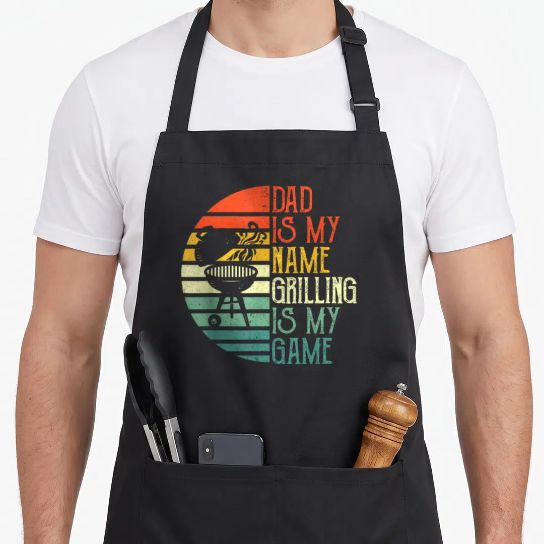 Game father day apron - Retro striped sun and grill with Dad Is My Name Grilling Is My Game text — shown on a black cotton canvas apron against a white background