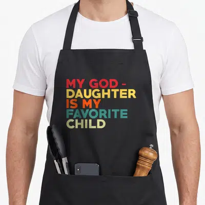 Goddaughter father day apron - Stacked colorful text reading MY GOD- DAUGHTER IS MY FAVORITE CHILD in red yellow orange — shown on a black cotton canvas apron against a white background