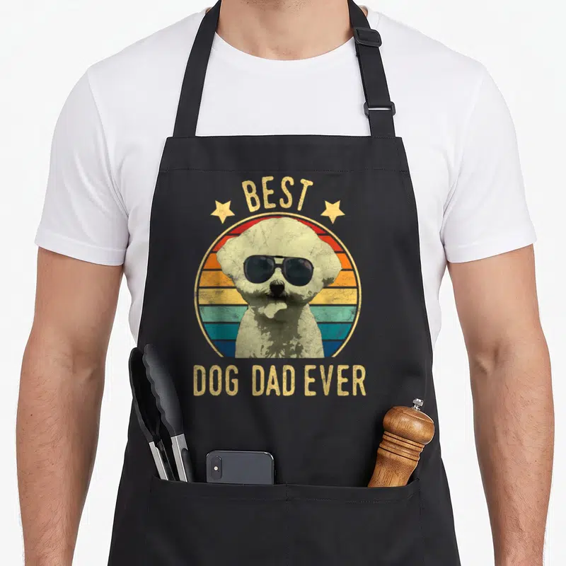 Bichon father day apron - Dog in sunglasses inside retro striped circle with BEST DOG DAD EVER text — shown on a black cotton canvas apron against a white background