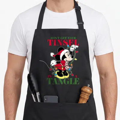 Tinsel holiday apron - Minnie Mouse tangled in Christmas lights with text DON'T GET YOUR TINSEL IN A TANGLE — shown on a black cotton canvas apron against a white background