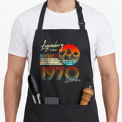 Legendary birthday apron - Retro sun with palm trees and stacked text Legendary since AUGUST 1970 Limited edition — shown on a black cotton canvas apron against a white background