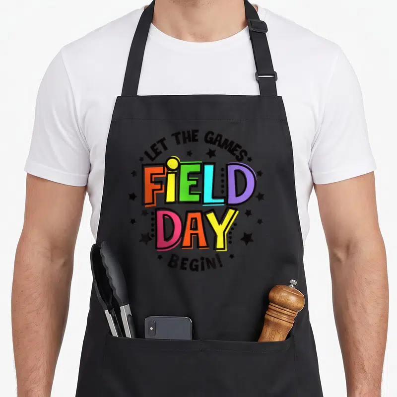 Field teacher apron - Multi-colored block text reading FIELD DAY surrounded by black stars and arched phrases — shown on a black cotton canvas apron against a white background