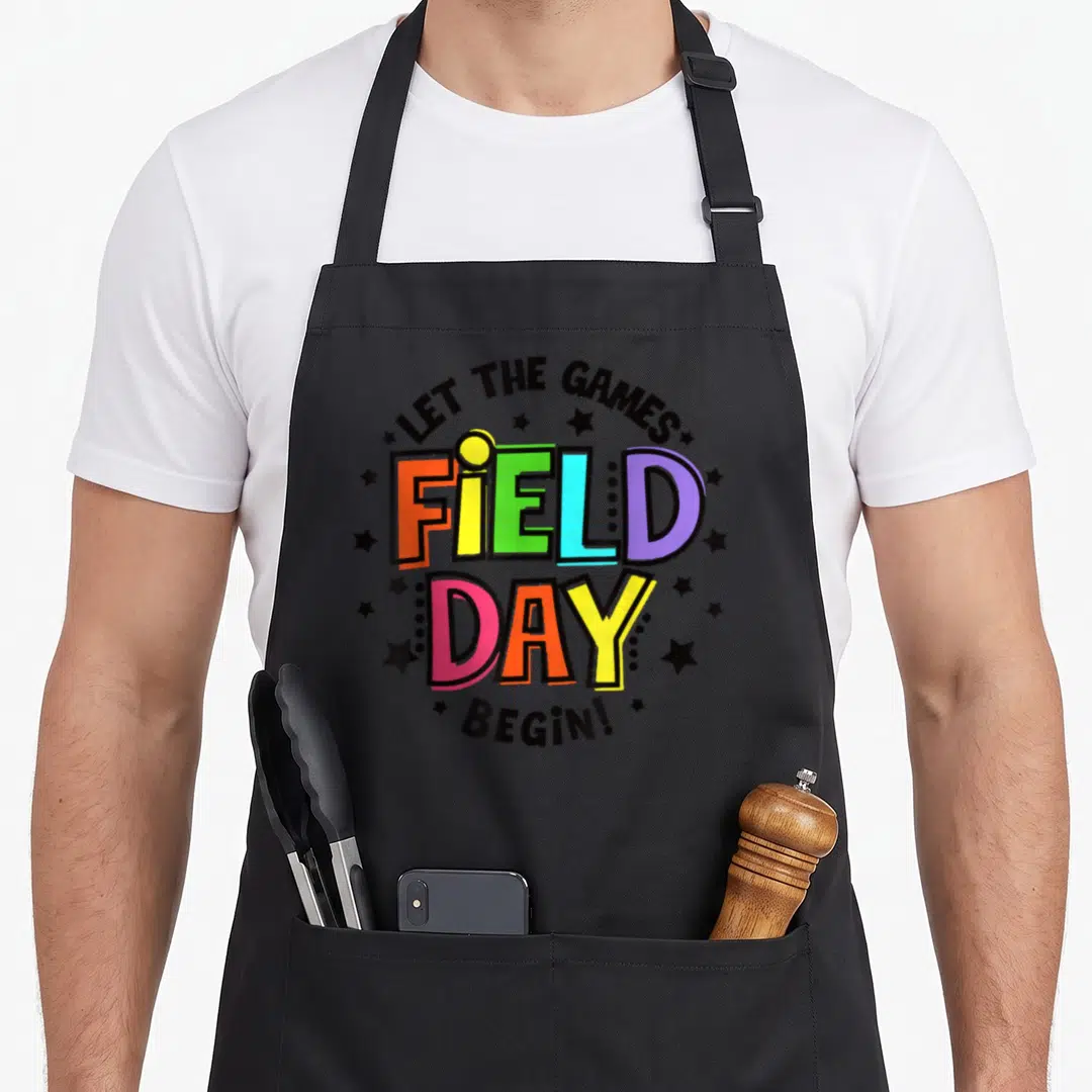 Field teacher apron - Multi-colored block text reading FIELD DAY surrounded by black stars and arched phrases — shown on a black cotton canvas apron against a white background