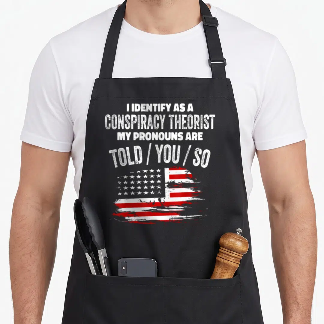 Conspiracy a's aprons - Distressed USA flag below white text reading I Identify As A Conspiracy Theorist My Pronouns — shown on a black cotton canvas apron against a white background