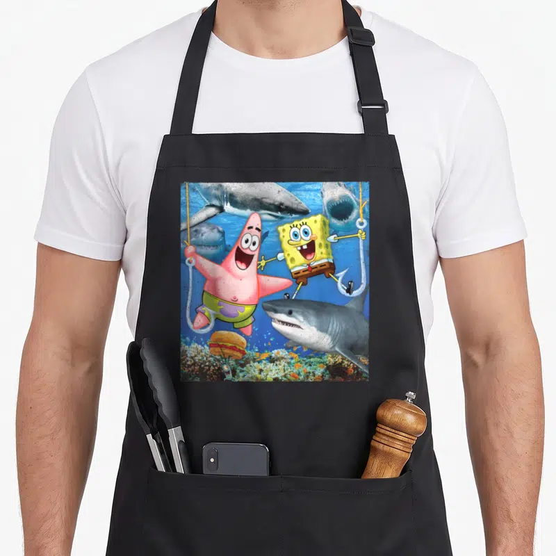 Perfect patrick apron - SpongeBob and Patrick hanging from hooks surrounded by grey sharks above a burger on coral — shown on a black cotton canvas apron against a white background