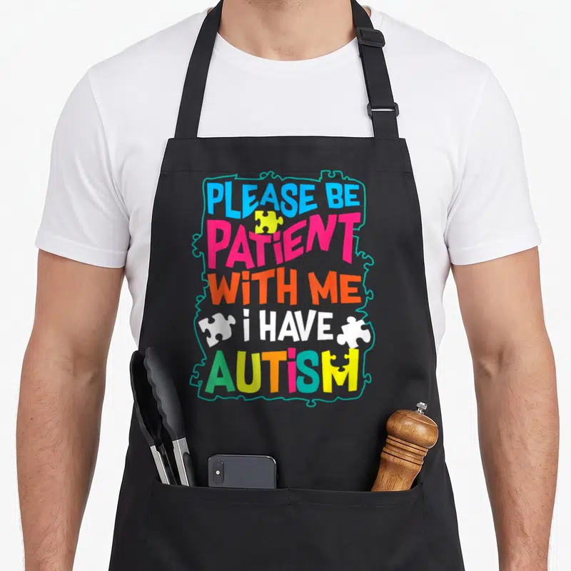 Patient glam aprons - Colorful text PLEASE BE PATIENT WITH ME I HAVE AUTISM with teal puzzle border — shown on a black cotton canvas apron against a white background