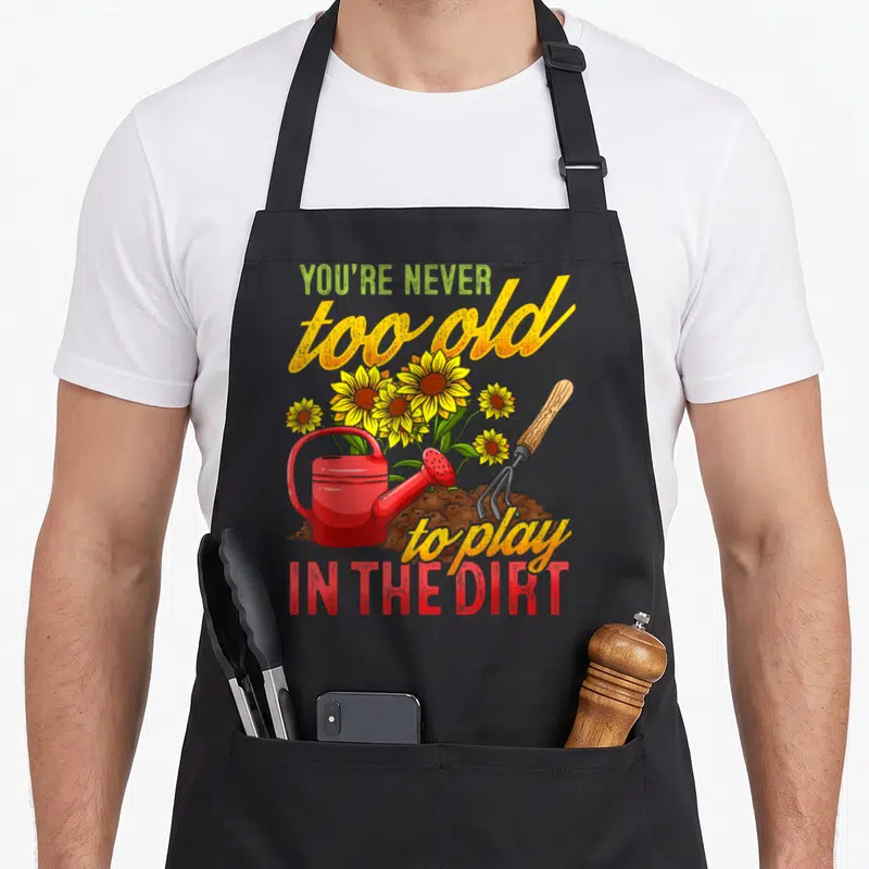Gardening plant gardener apron - Retro art showing red watering can, garden fork, and yellow sunflowers in brown dirt — shown on a black cotton canvas apron against a white background