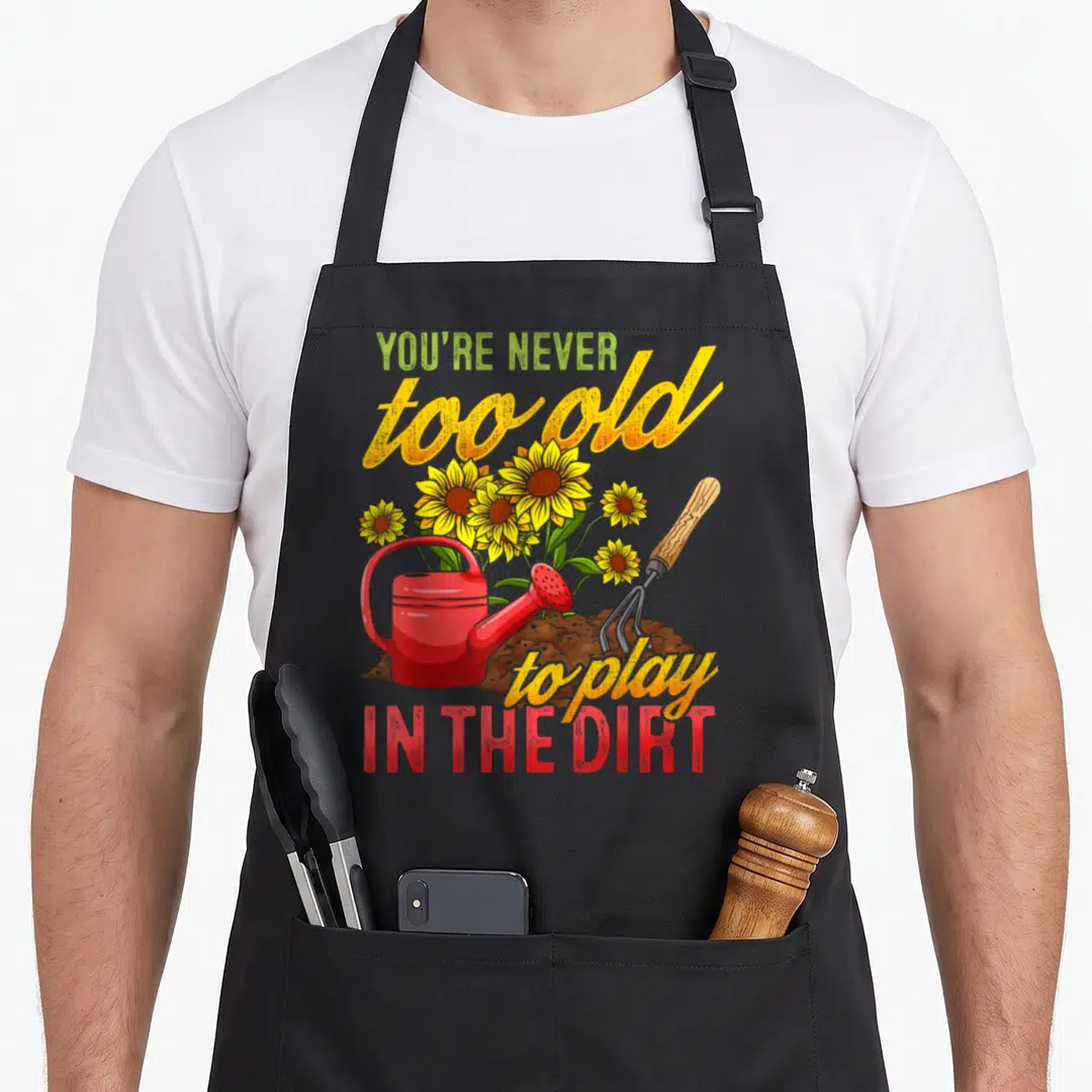Gardening plant gardener apron - Retro art showing red watering can, garden fork, and yellow sunflowers in brown dirt — shown on a black cotton canvas apron against a white background