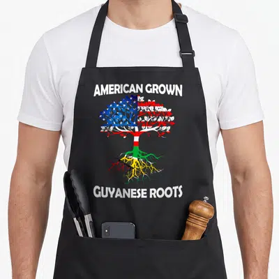 Grown guyana apron - Tree with US flag leaves and Guyanese roots flanked by American Grown Guyanese Roots text — shown on a black cotton canvas apron against a white background