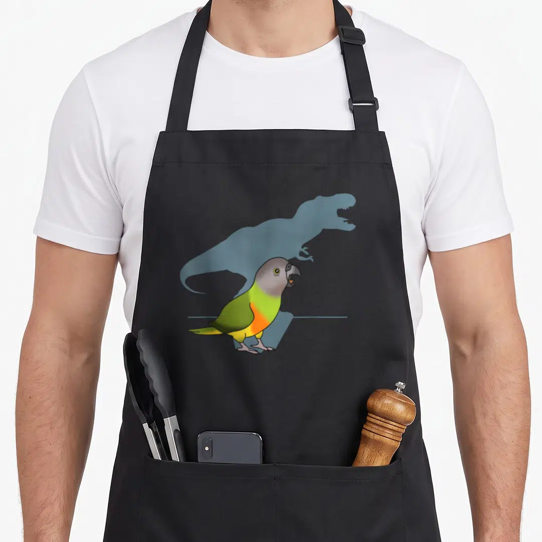 Senegal meme apron - Green and yellow parrot with blue dinosaur silhouette shadow in cartoon style — shown on a black cotton canvas apron against a white background