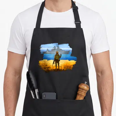 Stamp ukrainian aprons - Soldier on yellow ground facing grey warship in blue yellow brush stroke flag shape — shown on a black cotton canvas apron against a white background