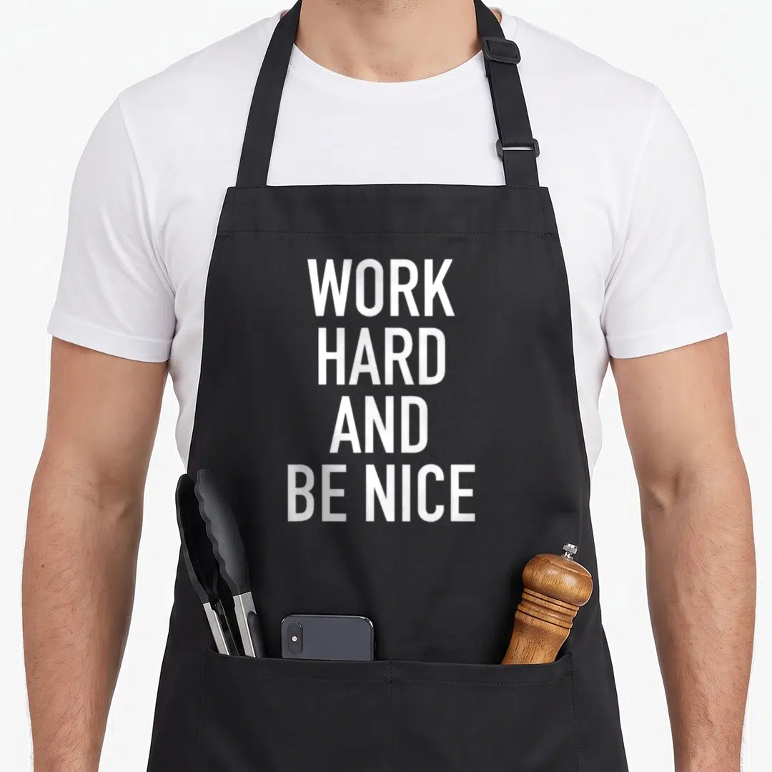 Funny motivational work hard apron - White stacked text reading WORK HARD AND BE NICE in bold font — shown on a black cotton canvas apron against a white background